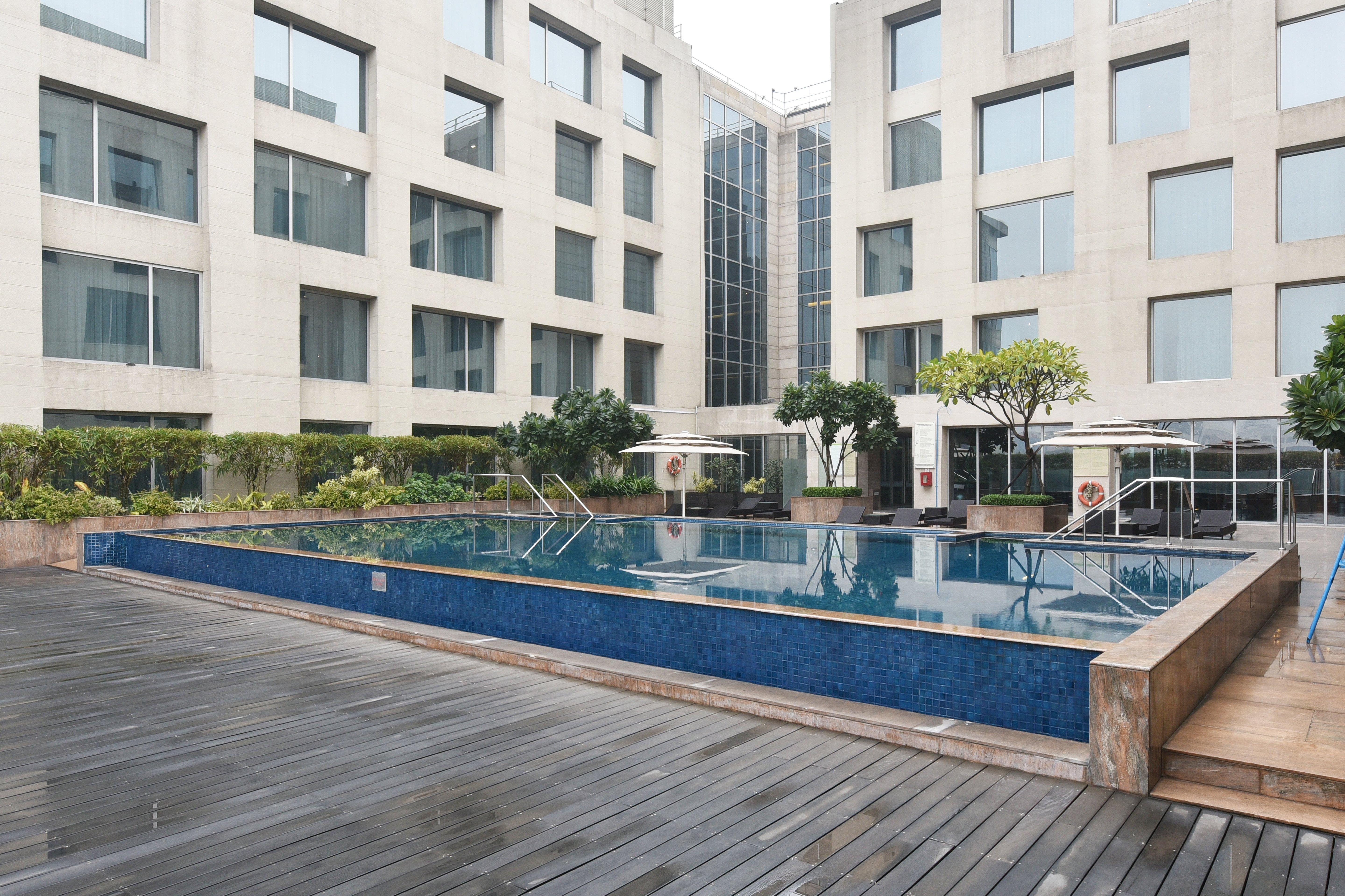 Holiday Inn New Delhi Aerocity By Ihg Екстериор снимка The photo shows a modern outdoor swimming pool situated in a courtyard surrounded by several buildings. The pool has a sleek design with a blue tiled edge and is bordered by wooden decking. There are a few trees and plants around the area, adding som