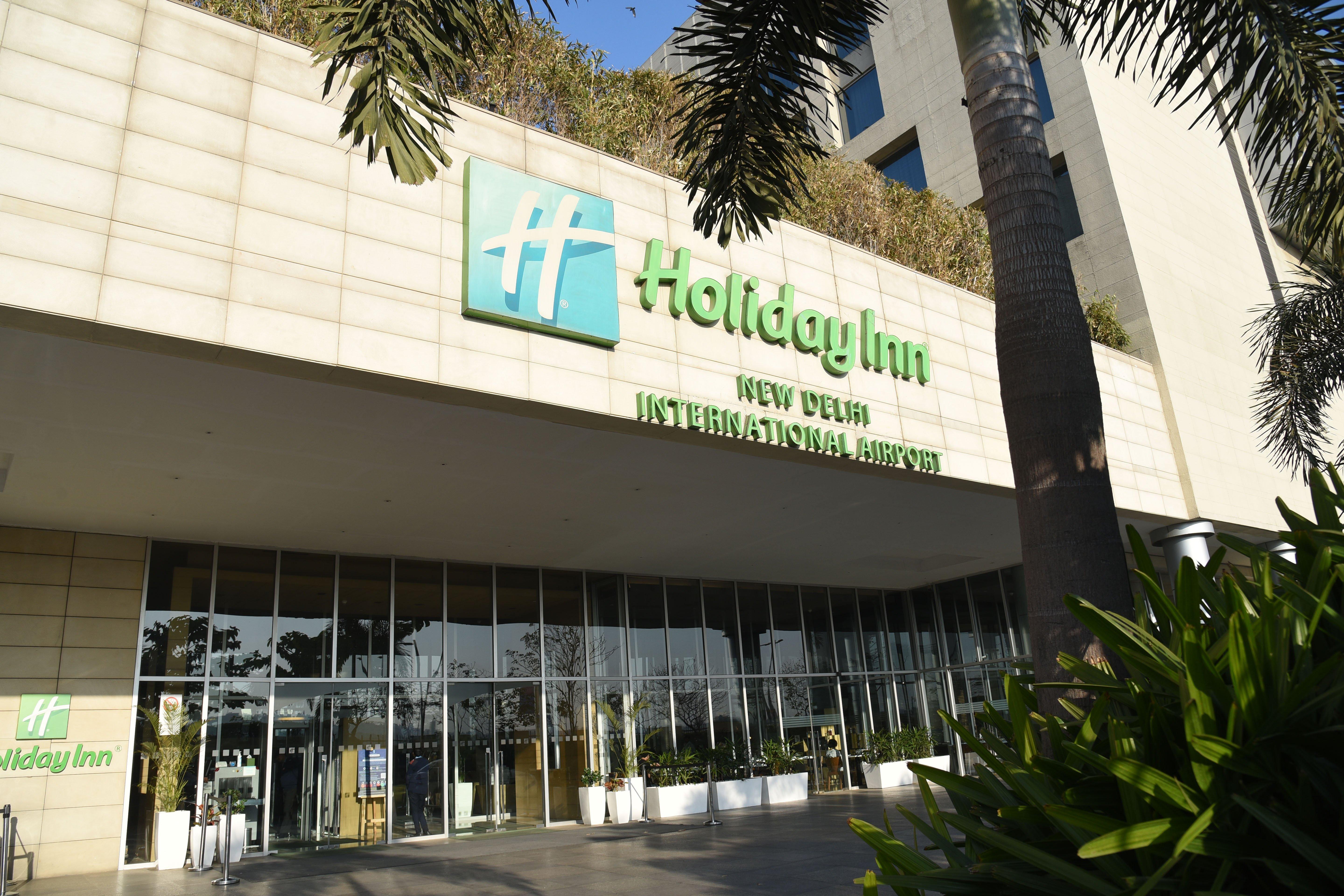 Holiday Inn New Delhi Aerocity By Ihg Екстериор снимка The photo shows the exterior of a Holiday Inn hotel, specifically the Holiday Inn New Delhi International Airport. The building features a modern design with a prominent green and white sign displaying the hotel name. There are large glass doors at t