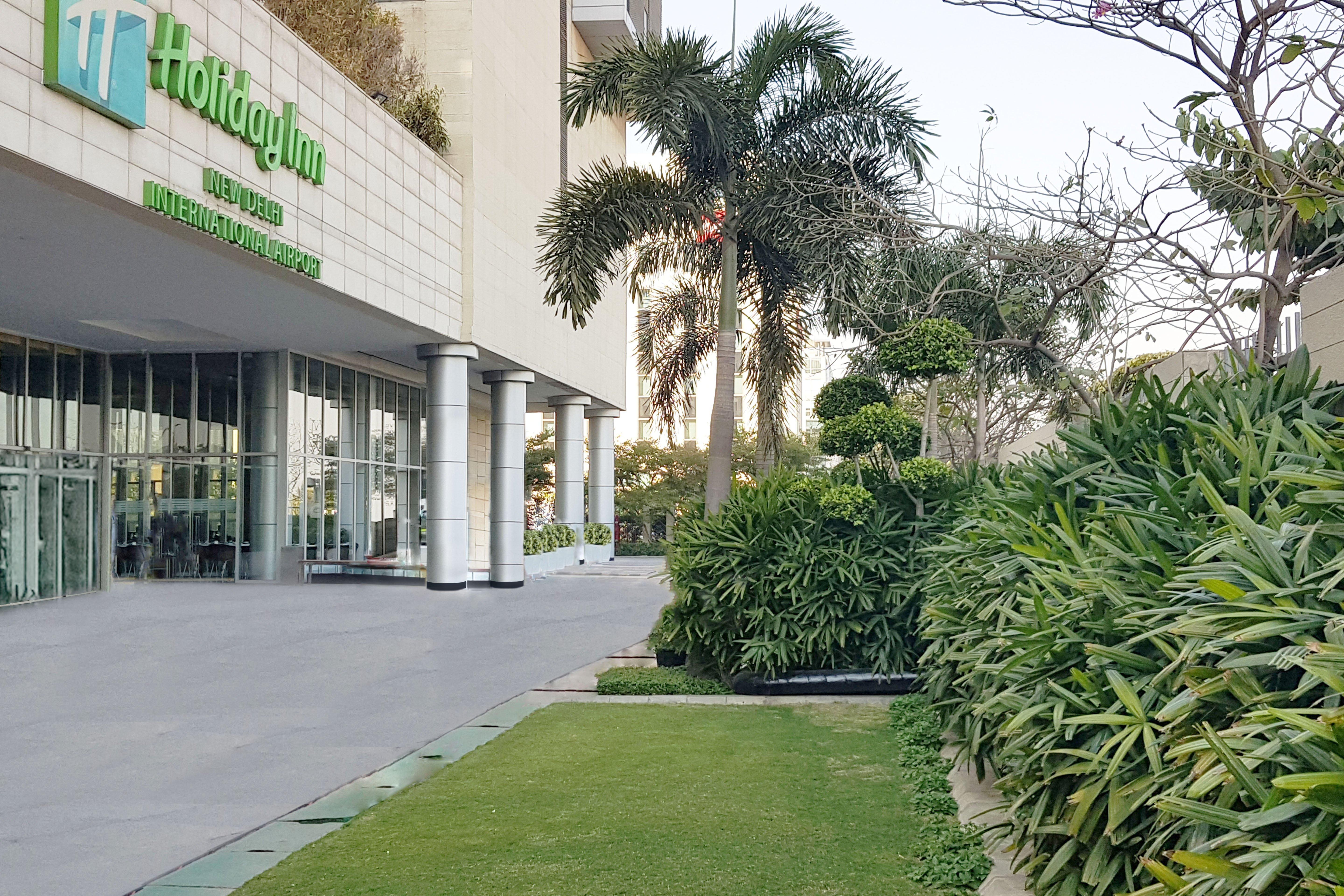 Holiday Inn New Delhi Aerocity By Ihg Екстериор снимка The photo shows the exterior of a Holiday Inn hotel. In the foreground, there is a well-maintained green lawn and various ornamental plants, creating a pleasant garden area. The building features large glass doors and columns, and the hotel's name an